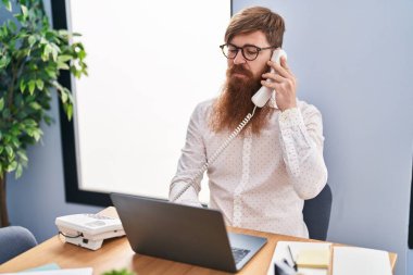 Young redhead man business worker using laptop talking on smartphone at office