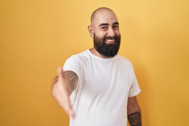 Sarı arka planda dikilen sakallı ve dövmeli genç İspanyol adam selamlaşma ve hoş geldin hediyesi olarak gülümsedi. başarılı iş. 