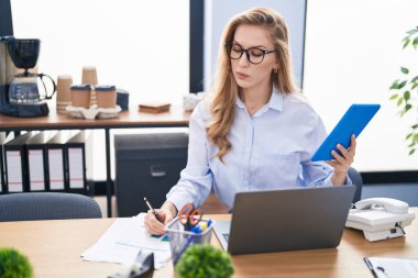 Ofiste çalışan genç sarışın iş kadını dizüstü bilgisayar ve dokunmatik ped kullanıyor.