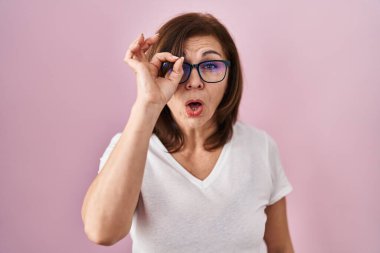 Middle age hispanic woman standing over pink background doing ok gesture shocked with surprised face, eye looking through fingers. unbelieving expression. 