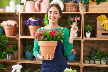 Young hispanic woman working at florist shop holding plant smiling happy pointing with hand and finger to the side 