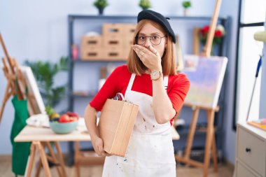 Sanat stüdyosundaki genç kızıl saçlı kadın sanat davasını tutarken ağzı yanlışlıkla ellerle kapatırken şok oldu. gizli kavram. 