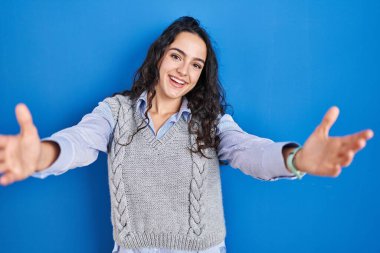 Genç esmer kadın mavi arka planda duruyor kameraya bakıyor kucaklaşmak için kollarını açmış gülümsüyor. Mutluluğu kucaklayan neşeli bir ifade.. 