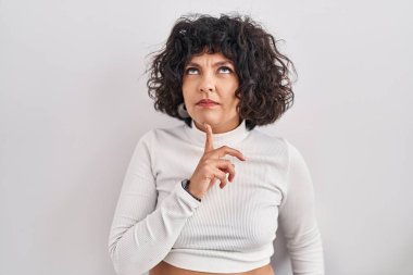 Kıvırcık saçlı İspanyol kadın izole edilmiş arka planda durup parmak çenesinde şüpheye yoğunlaşmış ve kafasını kaldırıp merak ediyor. 