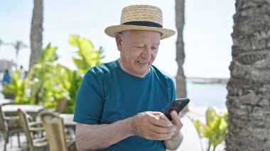 Deniz kenarında akıllı telefon kullanarak yaz şapkası takan yaşlı, kır saçlı bir turist.