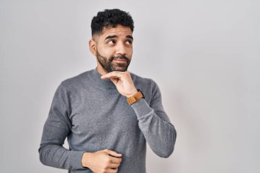 Hispanic man with beard standing over white background with hand on chin thinking about question, pensive expression. smiling with thoughtful face. doubt concept. 