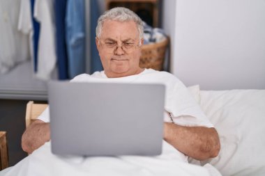 Yatak odasında dizüstü bilgisayar kullanan orta yaşlı, kır saçlı bir adam.