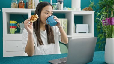 Genç, güzel, İspanyol bir kadın yemek odasında dizüstü bilgisayar kullanarak kahvaltı yapıyor.