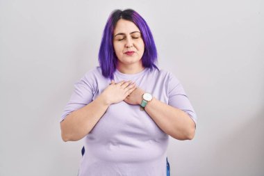 Artı cüsseli, mor saçlı bir kadın izole edilmiş arka planda duruyor. Elleri göğsünde, gözleri kapalı ve yüzünde minnet dolu bir jest ile gülümsüyor. sağlık kavramı. 