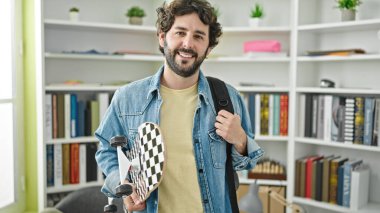 Kütüphanede kaykay tutan sırt çantası takan İspanyol bir öğrenci.