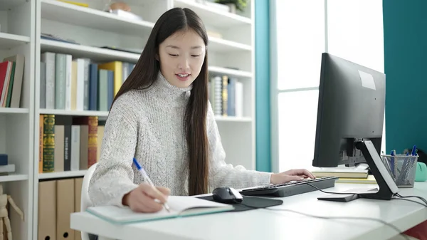 Young chinese woman student using computer writing on notebook at library university