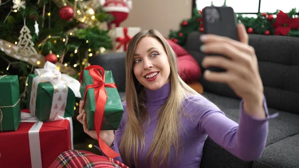 年轻的金发女人坐在家里的圣诞树旁 用智能手机自拍 — 图库照片