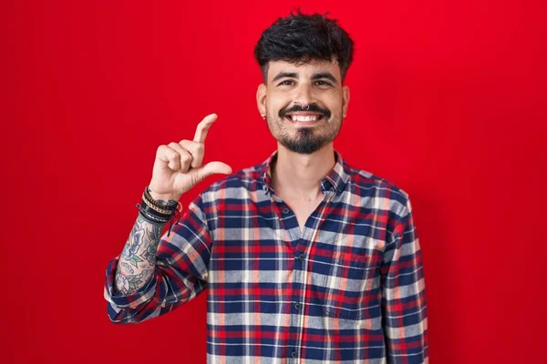 赤い背景の上に髭を生やした若いヒスパニック系の男が笑顔で 指を見てカメラで小さなサイズの看板をして手で自信を持ってジェスチャー 概念を測定し — ストック写真