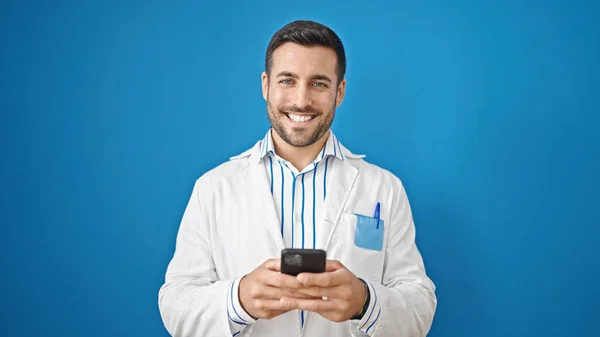 Νεαρός Ισπανόφωνος Γιατρός Χαμογελά Χρησιμοποιώντας Smartphone Απομονωμένο Μπλε Φόντο — Φωτογραφία Αρχείου