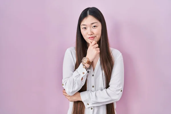 カメラを見て自信を持ってピンクの背景に立っている中国の若い女性が交差腕と手であごに提起笑みを浮かべて ポジティブな — ストック写真