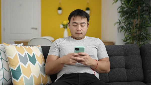 Young chinese man using smartphone sitting on sofa at home
