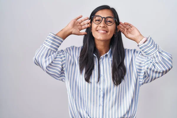 若いヒスパニック系の女性の眼鏡を指で耳を引く笑顔を着て 面白いジェスチャー オーディション問題 — ストック写真