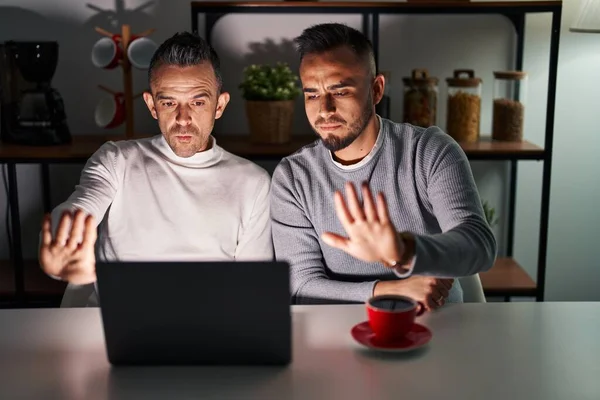 Homosexual couple using computer laptop doing stop sing with palm of the hand. warning expression with negative and serious gesture on the face.