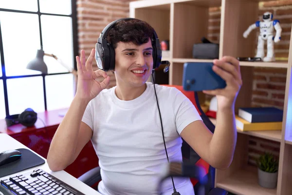 若いです非バイナリの男は指でOkサインを行うスマートフォンでビデオゲームをプレイします — ストック写真