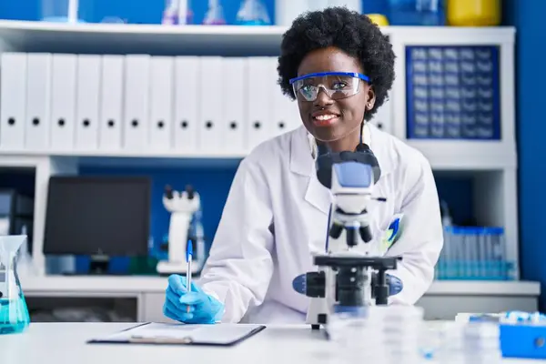 若いアフリカ系アメリカ人女性科学者が研究室で顕微鏡を使って文書を書く — ストック写真