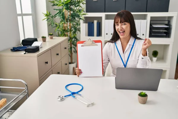 Young Brunette Doctor Woman Holding Clipboard Screaming Proud Celebrating Victory — Stock Photo, Image