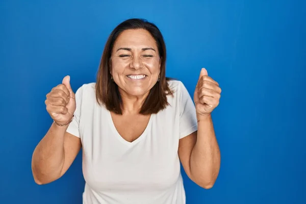 腕を上げて成功のために興奮した青い背景の上に立つヒスパニック系の成熟した女性と目は笑顔勝利を祝う閉じました 勝者のコンセプト — ストック写真