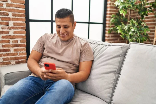 年轻的拉丁男人坐在家里的沙发上用智能手机 — 图库照片