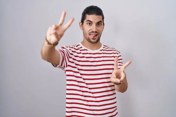 一个长头发的西班牙男人站在孤立的背景上 用舌头笑着伸出双手的手指做胜利的标志 第二点 — 图库照片