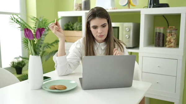 年轻美丽的惊慌失措的女人在饭厅用笔记本电脑吃饼干 — 图库照片