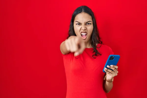 Młoda Brazylijka Pomocą Smartfona Czerwonym Tle Wskazując Niezadowolony Sfrustrowany Kamery — Zdjęcie stockowe
