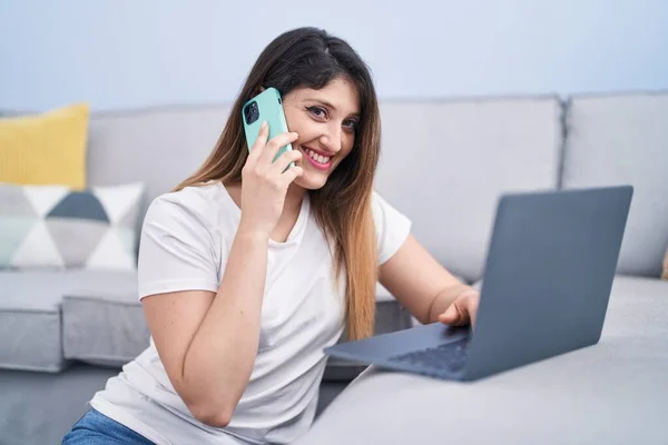 若いヒスパニック系の女性は自宅でスマートフォンで話してノートパソコンを使用して — ストック写真