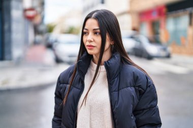 Young beautiful hispanic woman looking to the side with relaxed expression at street