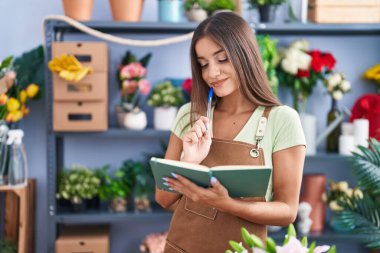 Young beautiful hispanic woman florist smiling confident reading notebook at flower shop