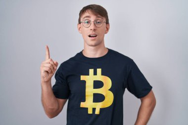 Bitcoin tişörtlü beyaz sarışın adam başarılı bir fikir olduğunu işaret ediyor. Heyecanlı ve mutlu. Bir numara.. 