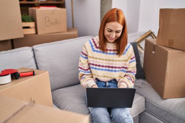 Beyaz kadın yeni evinde dizüstü bilgisayar kullanarak gülümsüyor.