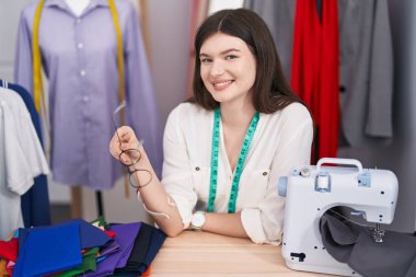 Young caucasian woman tailor smiling confident sitting on table at tailor shop