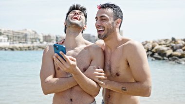 İki erkek turist kumsalda akıllı telefon kullanarak gülümsüyor.
