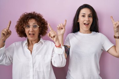 İspanyol anne ve kızı pembe arka planda gündelik beyaz tişört giyip şaşkın şaşkın gülüyor, parmaklarıyla işaret ediyor ve kollarını kaldırıyordu.. 