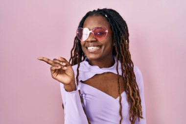 Pembe arka planda örülü saçları olan Afrikalı bir kadın yüzünde kocaman bir gülümsemeyle kameraya el ve parmak ile bakıyor.. 