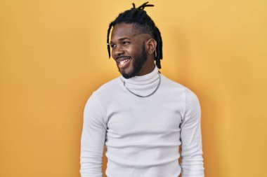 Sarı arka planda balıkçı yaka kazak giyen Afrikalı bir adam yüzünde gülümseme, doğal bir ifade ile yan yana bakıyor. Kendinden emin gülen. 