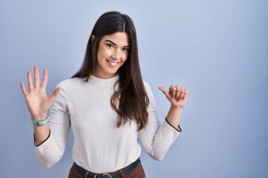 Genç esmer kadın mavi arka planda duruyor. Altıncı parmaklarını gösterip işaret ediyor. Kendinden emin ve mutlu bir şekilde gülümsüyor.. 