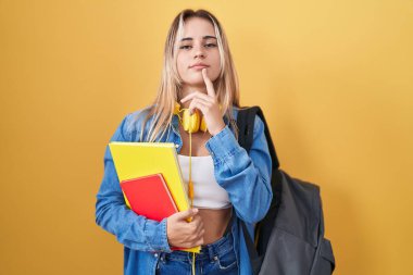Sırt çantası takan ve elinde kitap tutan genç sarışın kadın şüpheye odaklanmış, çenesinde parmak olan ve kafasını kaldırıp merak eden. 