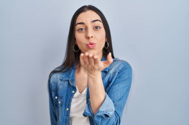 Latin bir kadın mavi arka planda dikilip kameraya bakarken havada bir öpücük savuruyor ve sevimli ve seksi oluyor. aşk ifadesi. 