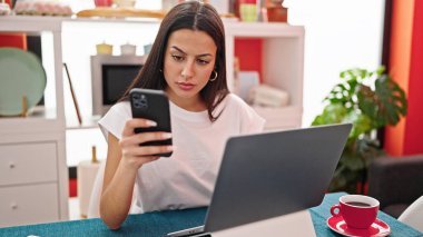 Yemek odasında dizüstü bilgisayar ve akıllı telefon kullanan güzel bir İspanyol kadın.