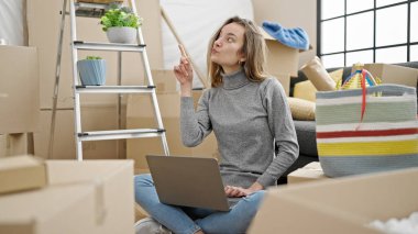 Genç beyaz kadın yeni evinde yerde otururken dizüstü bilgisayar kullanmayı düşünüyor.