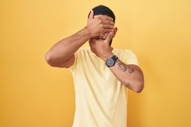 Young hispanic man standing over yellow background covering eyes and mouth with hands, surprised and shocked. hiding emotion 