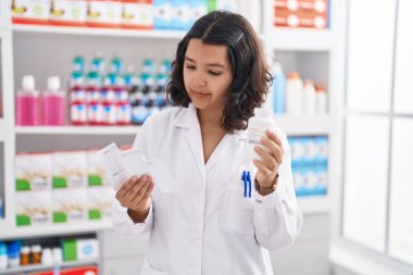 Young woman pharmacist holding pills bottle reading prescription at pharmacy