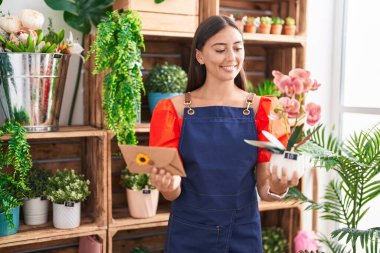 Young beautiful hispanic woman florist holding envelope letter and plant at florist