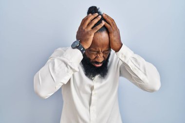 Afro-Amerikalı bir adam mavi arka planda dikiliyor. Baş ağrısı, çaresiz ve stresli. Çünkü acı ve migren. Eller başının üstünde.. 