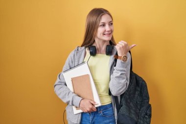 Sırt çantası takan, elinde kitaplarla gülümseyen, gülümseyen ve baş parmağıyla yüzünü gösteren beyaz bir kadın.. 